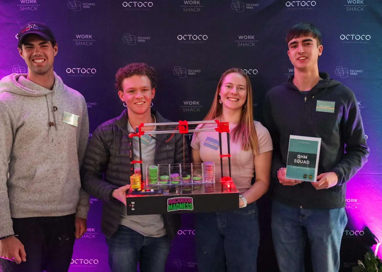 The winning Ohm Squad team standing with their gesture-controlled sorting machine under purple stage lighting, in front of an Octoco and Workshack backdrop
